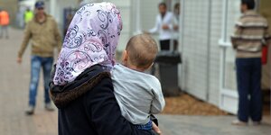 Ein Frau mit Kopftuch trägt ein Kleinkind durch eine Flüchtlingsunterkunft.