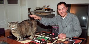 Akif Pirinçci am Schreibtisch sitzend. Er präsentiert eines seiner Bücher und streichelt eine Katze, die sich auf dem Tisch befindet.