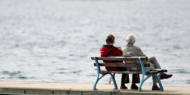 Zwei Rentner_innen sitzen auf einer Bank vor einem See