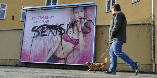 Auf einem mit „Sexismus“ beschmierten Plakat ist eine Frau in Dessous zu sehen. Ein Mann spaziert vorbei