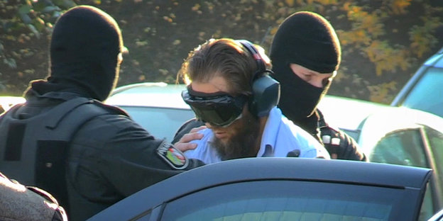 Zwei Polizisten schieben einen Mann mit dunkler Brille und Kopfhörern in ein Auto