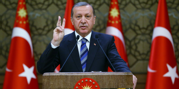 Der türkische Präsident Erdoğan steht an einem Pult und hebt drei Finger