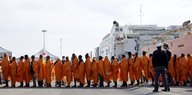 Männer mit orangenen Umhängen stehen in einer Schlange. Hinter ihnen ist ein Schiff zu sehen