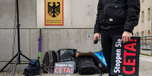 Einige Protestutensilien, ein Lautsprecherständer und ein mensch mit einem Anti-Ceta-Schild stehen vor einer grauen Mauer