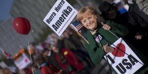 ein Demonstrant mit Merkel-Maske und zwei Schildern, auf einem steht „Hurra Hurra Amerika“, auf dem anderen „Ich liebe USA“
