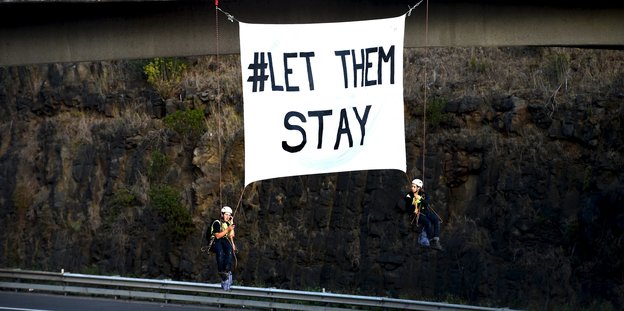 Zwei Menschen in Kletterausrüstung hängen mit einem Transparent auf dem „Let them stay“ steht von einer Brücke