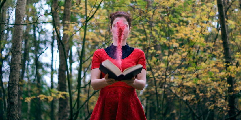 Eine Frau im roten Kleid steht im Wald und liest ein Buch, aus dem rote Farbe dampft