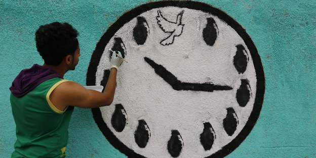 ein junger Mann malt eine großes Ziffernblatt an eine Wand, statt Zahlen sind Handgranaten zu sehen und oben in der Mitte eine Friedenstaube