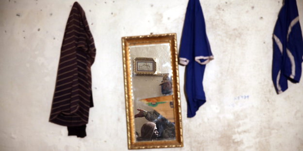 In einem Spiegel an einer Wand sieht man einen libyschen Soldaten und einen Flüchtling. Daneben hängen dunkle T-Shirts