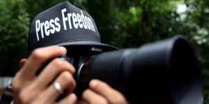 Ein Fotograf hält eine Kamera vor das Gesicht und trägt ein Cap mit der Aufschrift "Press Freedom"