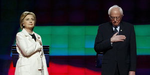 Clinton und Sanders stehen mit ernster Miene und Hand auf dem Herzen auf der Bühne