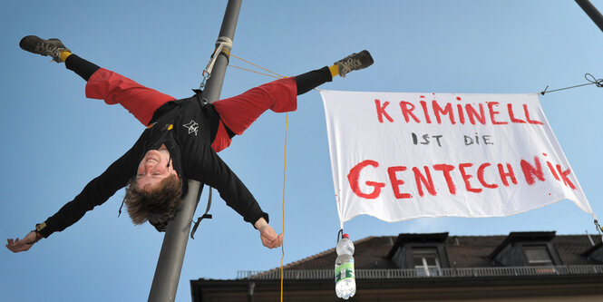 Eine Frau hängt kopfüber an einer Stange, dahinter ein Banner gegen Gentechnik