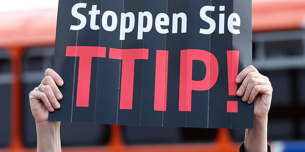 Zwei Hände halten ein schwarzes Schild mit der Aufschrift „Stoppen Sie TTIP!“ in die Luft