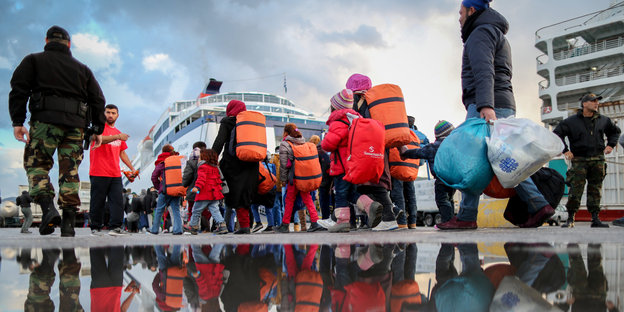 Mehrere Menschen mit Gepäck gehen auf ein Schiff zu, sie spiegeln sich in einer Pfütze