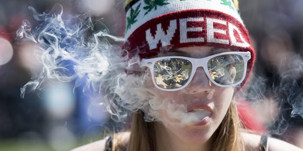 Eine junge Frau mit Sonnenbrille und Wollmütze, auf der "Weed" steht, atmet Rauch aus