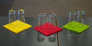 Drei Stapel Servietten liegen auf einem Tisch. Sie sind gelb, rot und grün. Darauf stehen umgedrehte Gläser