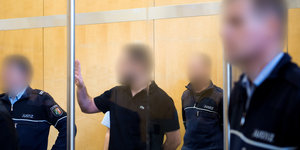 Ein wegen Unterstützung terroristischer Vereinigungen angeklagte Mann steht im Oberlandesgericht Düsseldorf neben Justizbeamten hinter einer Glasscheibe im Gerichtssaal.
