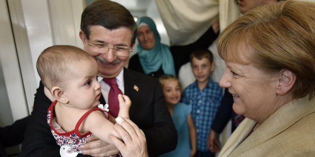 Davutoglu hält ein Baby im Arm, Merkel lächelt dazu