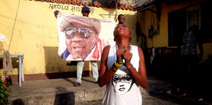 Eine junge afrikanische Frau trauert in einem Hof, hinter ihr hält ein junger Mann ein Transparent mit Papa Wembas Gesicht in die Höhe
