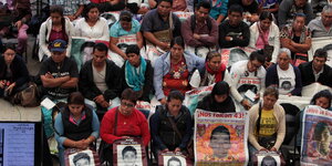 Menschen sitzen in Reihen hintereinander. Sie halten Postern mit den Gesichtern verschwundener Studenten in den Händen