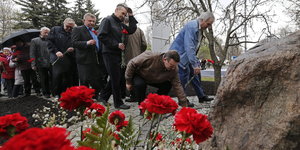 Menschen stehen vor einem Stein in einer Reihe und legen rote Rosen nieder