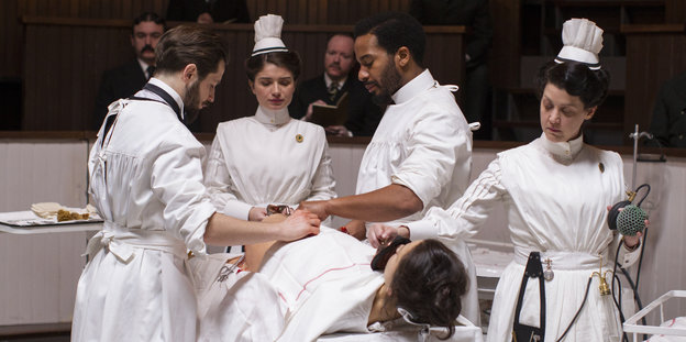 Ärzte und Krankenschwestern in historischer Kleidung stehen um einen Operationstisch, auf dem ein Patient liegt
