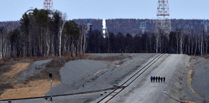 einige Menschen laufen neben Gleisen, im Hintergrund eine Rakete und Wald
