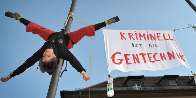 Eine Frau hängt kopfüber an einem Fahnenmast neben einem Banner mit der Aufschrift "Kriminell ist die Gentechnik"