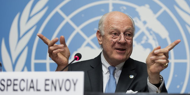 ein Mann deutet mit den Zeigefingern nach links und rechts, im Hintergrund das UN-Logo