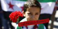 Ein Kind mit Nelke und dem Spruchband „Aleppo is burning“ vor einer syrischen Flagge