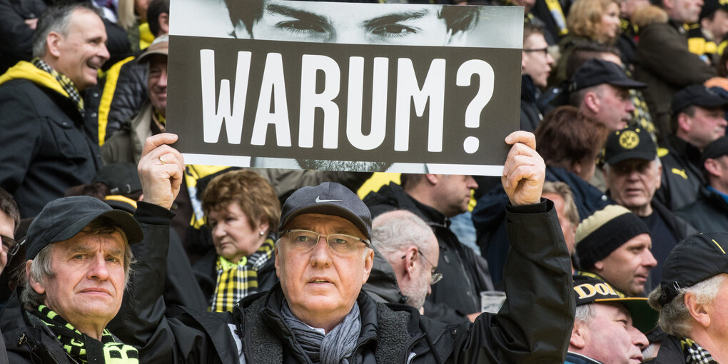 Ein Fän hält ein Schild hoch, auf dem "Warum?" steht