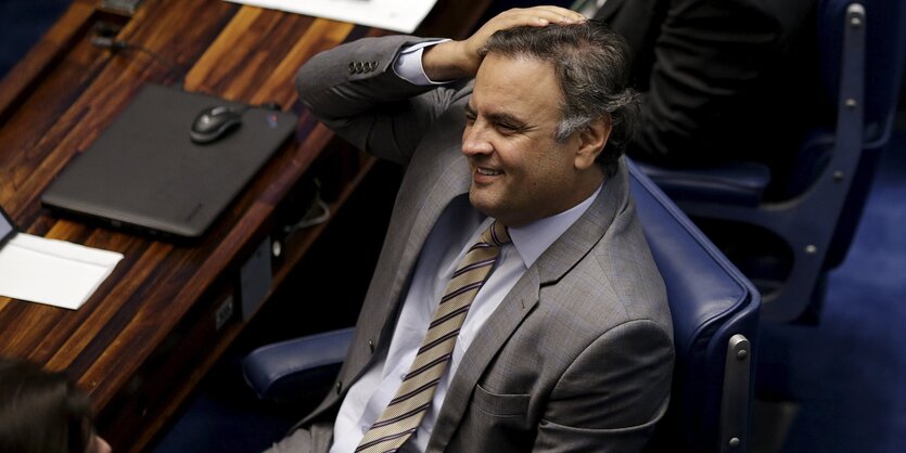 Aécio Neves sitzt lächelnd hinter einem Tisch und legt die Hand an den Kopf