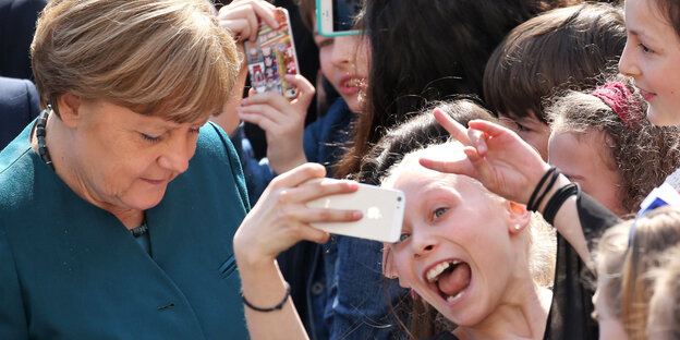 Merkel guckt nach unten, vor ihr eine Schülerin, die irr in ein zum Selfie gezücktes Handy grinst