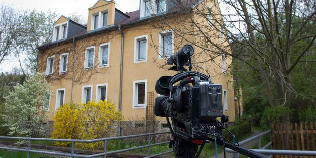 Vor einem braunen Haus steht eine Kamera auf einem Stativ