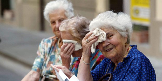 Drei alte Frauen nebeneinander, zwei trocknen sich die Stirn mit Tüchern