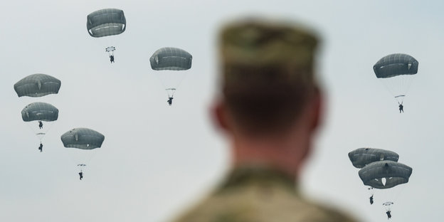 Ein Soldat blickt militärischen Fallschirmspringern entgegen, die im Landeanflug sind.