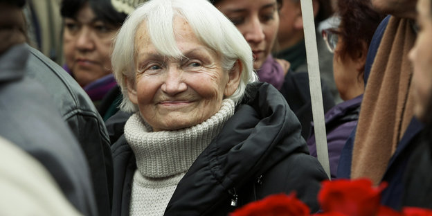 Margot Honecker steht in einer Menschenmenge, vor ihr hält jemand Nelken
