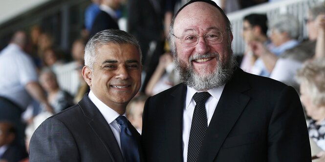 Auf der vollen Zuschauertribüne eines Stadions stehen zwei Männer in Anzug nebeneinander: Links Sadiq Khan und rechts der Rabbiner Ephraim Mirvis, der eine Kippa trägt.