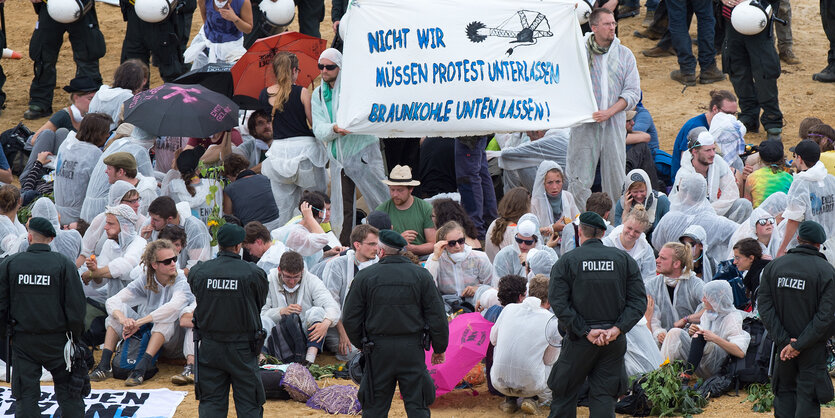 Mehrere Menschen sitzen in weißen Mond-Anzügen auf dem Boden, halten ein Transparent hoch und werden von Polizisten umkreist.