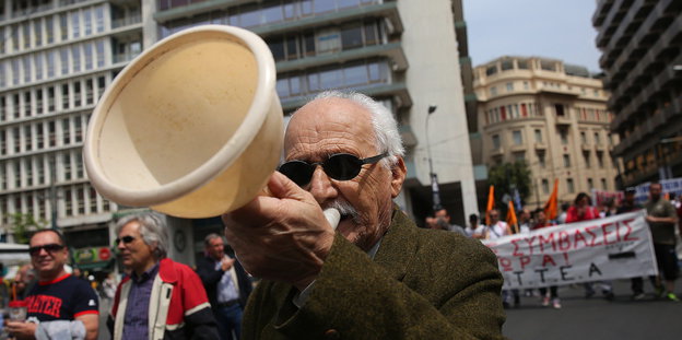 Ein Mann mit weißen Haaren und Sonnenbrille hält ein hautfarbenes Megaphon am Mund