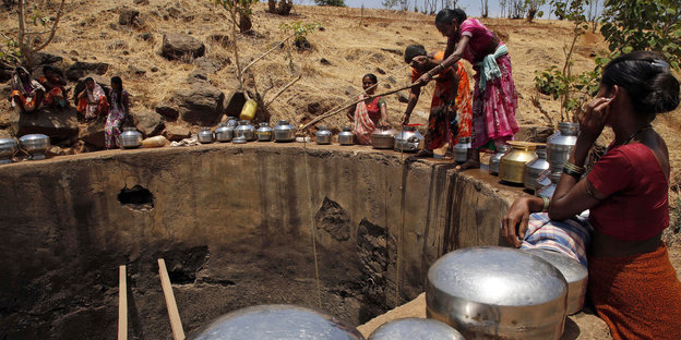 Frauen stehen mit Gefäßen am Rande eines Brunnens