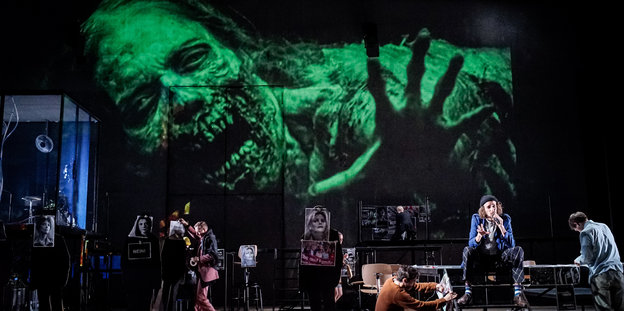 Eine Theaterbühne, auf der Menschen stehen, im Hintergrund eine grün-schwarze Projektion eines gruseligen menschlichen Monsters