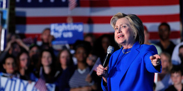 Hillary Clinton in blauem Anzug vor Anhängern und einer US-Flagge