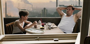 Zwei Menschen sitzen auf einem Balkon, im Hintergrund ein Industriegebiet
