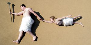 Ein Mann mit Fellhose und Steinhammer zieht eine Frau hinter sich her, beide liegen auf Sand