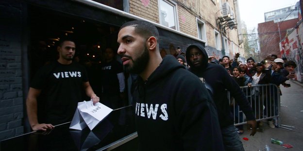 Drake trägt ein schwarzes Sweatshirt mit der Aufschrift „Views“ und läuft an einer Schlange wartender Leute vor einem Club vorbei