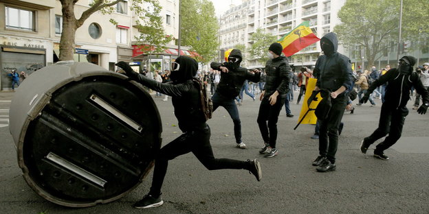 Schwarz gekleidet und vermummte Demonstranten rollen einen Altglascontainer die Straße entlang