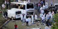 Ein zerstörter Bus und ein beschädigter Kleintransporter stehen nebeneinander, rund herum viele Menschen, teils in weißen Anzügen