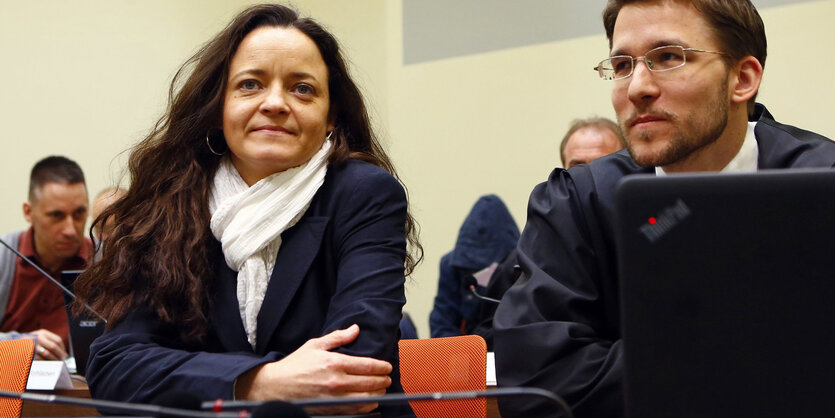 Eine Frau mit langen dunklen Haaren und weißem Schal sitzt neben einem Mann mit Brille und Robe