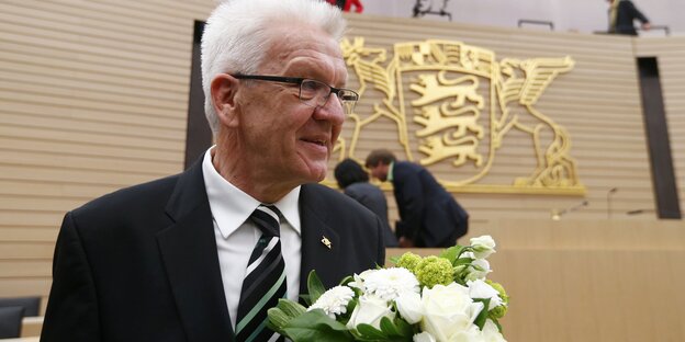Ein älterer Mann mit weiß-grauen Haaren trägt Anzug und eine grün-schwarz-gestreifte Krawatte, er hält einen weißen Blumenstrauß.